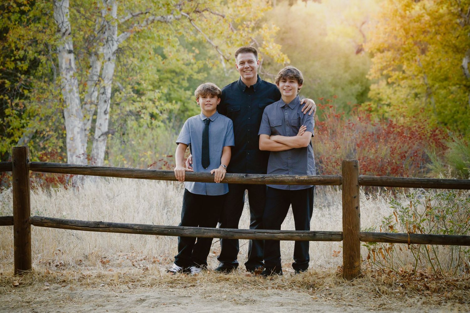 Man and two teen boys in dark shirts pose together behind a rustic wooden fence with golden autumn trees in the background.