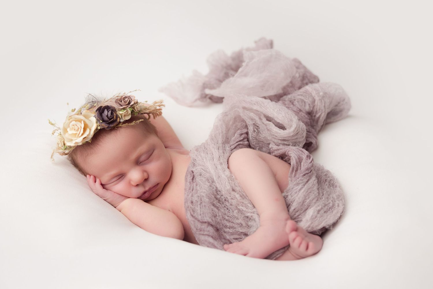 Newborn baby sleeping peacefully on white backdrop wearing floral headband and soft gray wrap.