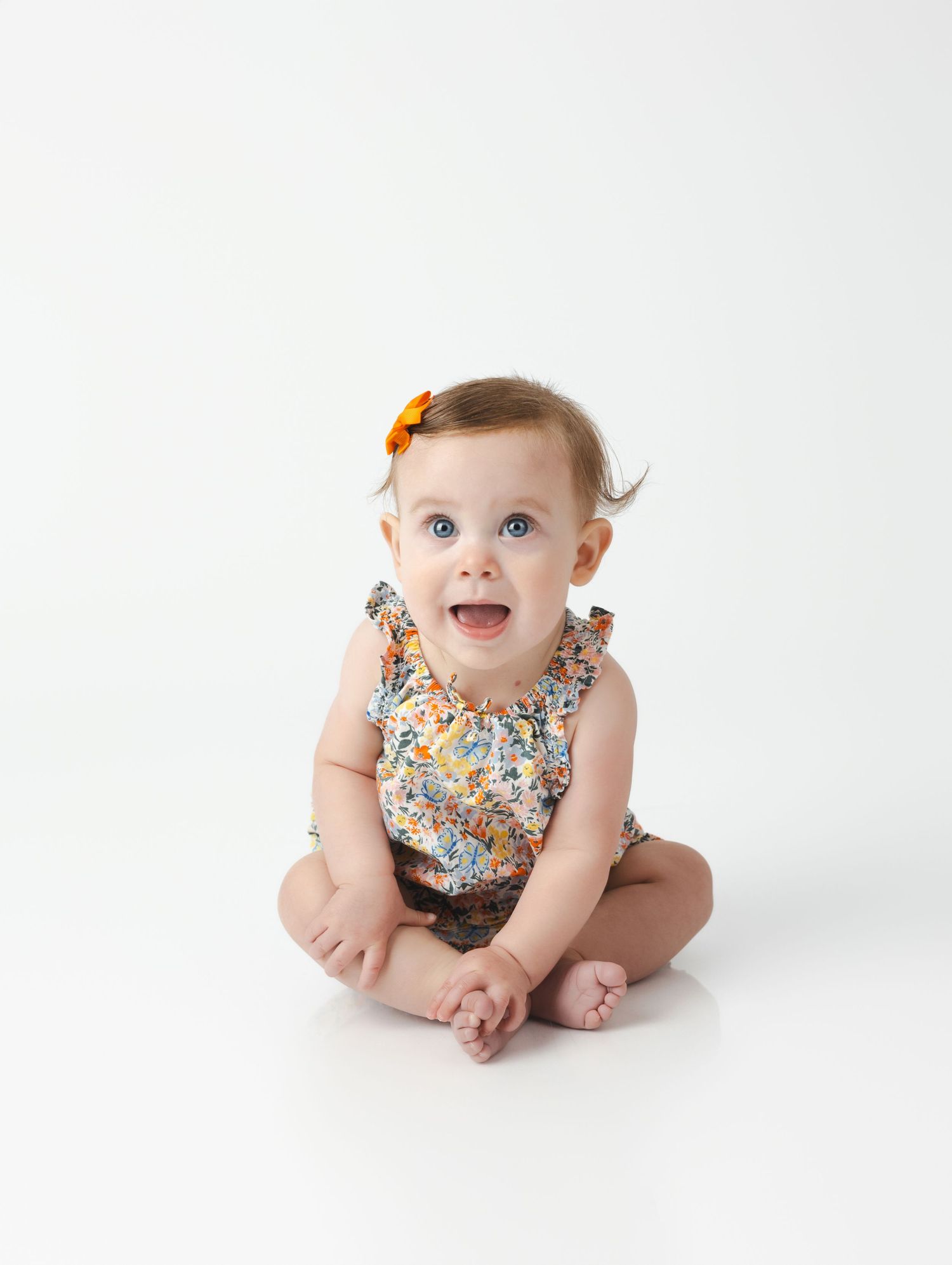 A baby wearing a floral print dress with orange hair clip sits on white background with a joyful expression.