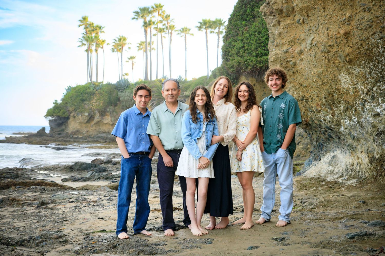 A group poses together on a rocky beach with tall palm trees and cliffs in the scenic coastal background.