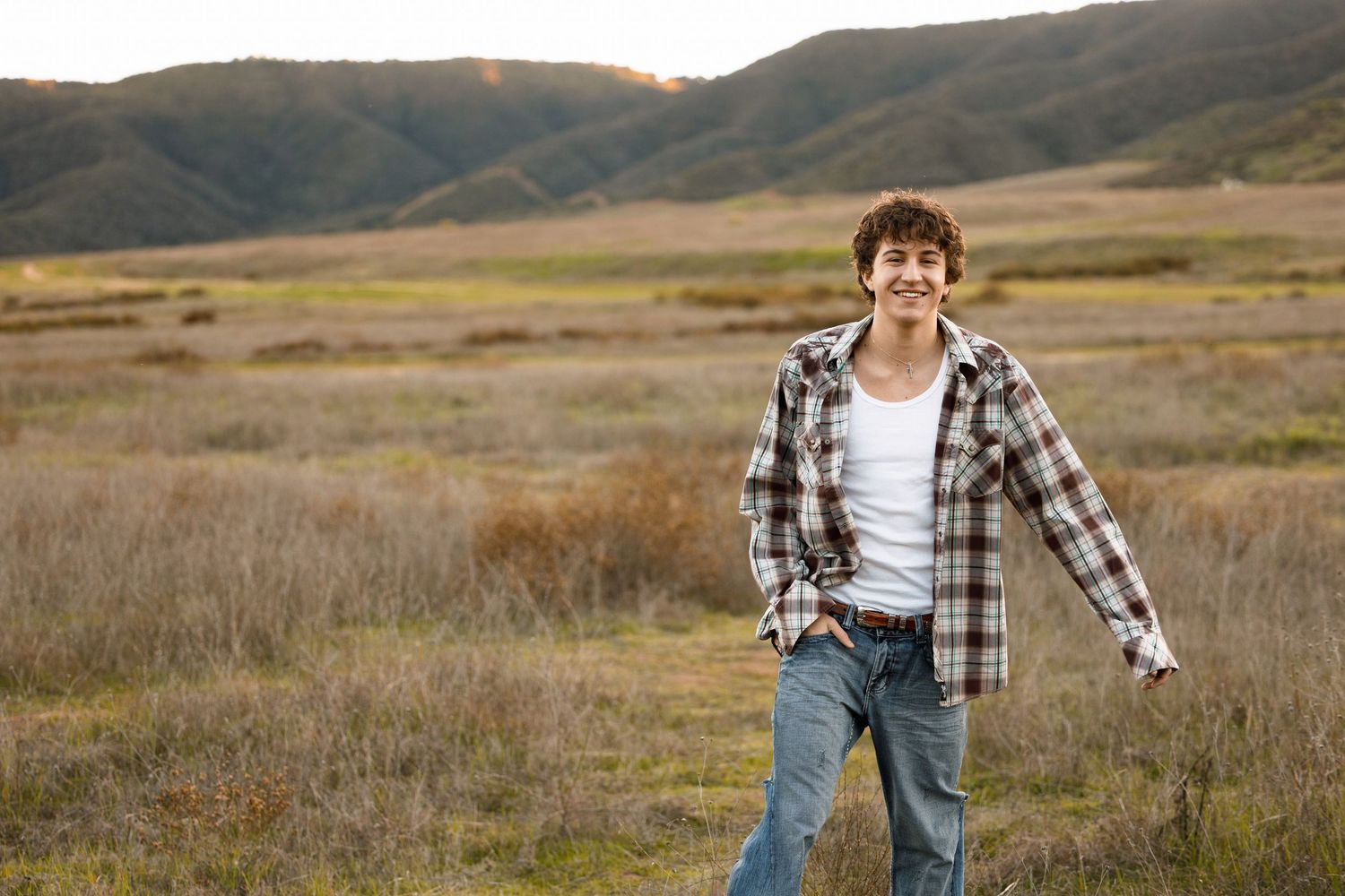 Person in plaid shirt and jeans walks through golden autumn field with mountains in background.