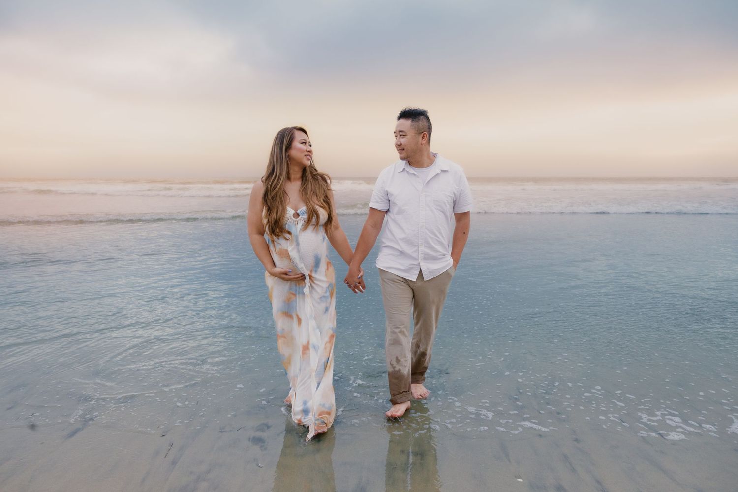 Couple holding hands walking on beach at sunset, woman in floral maxi dress, man in white shirt and khaki pants.