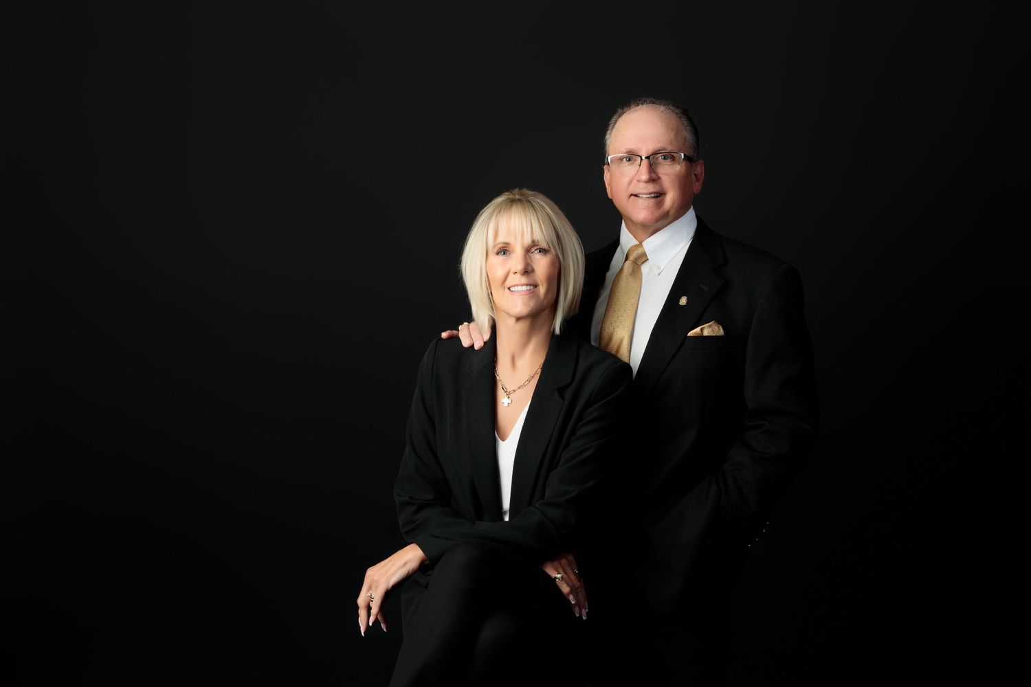 Elegant couple in formal attire poses for a professional portrait against a dark background.