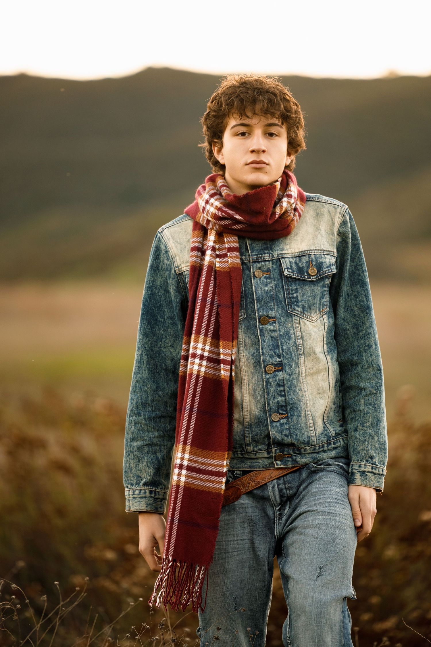 A person in denim jacket and plaid scarf sits in a field during golden hour lighting.