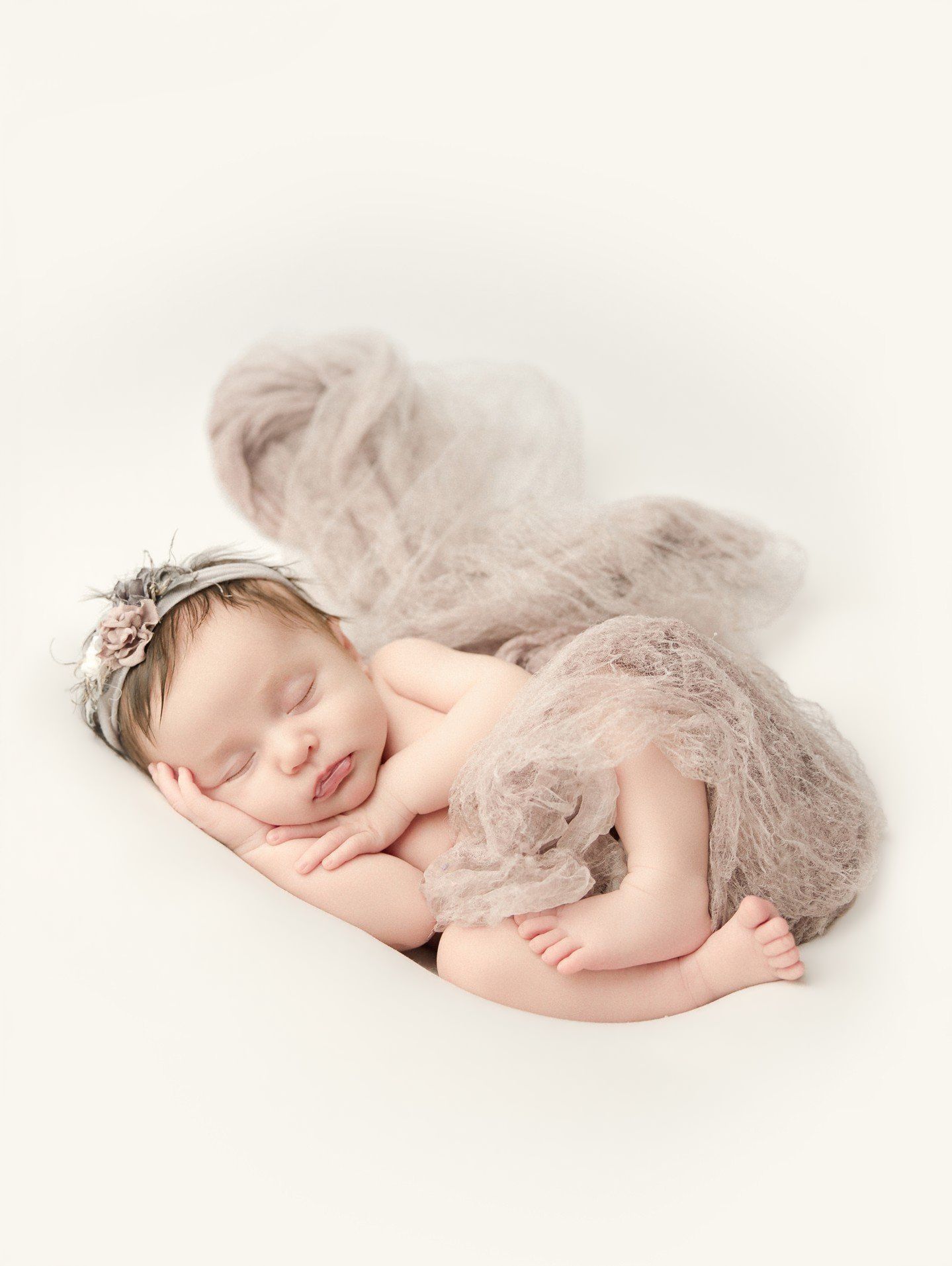 Peaceful newborn baby sleeping posed in soft lace wrap with floral headband on white background.