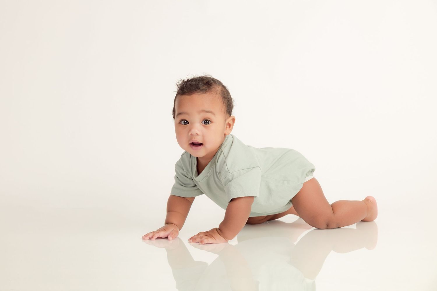 A curious baby in a green onesie crawls on a white floor, looking up with wide eyes.