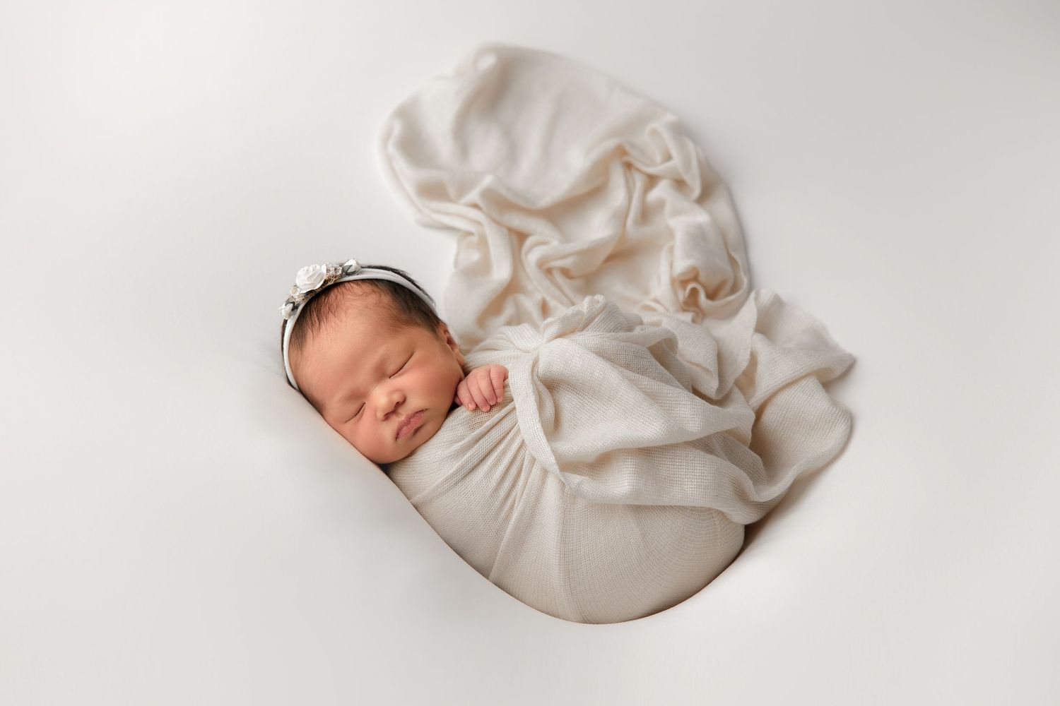 A sleeping newborn baby lies peacefully wrapped in soft white fabric against a light background.