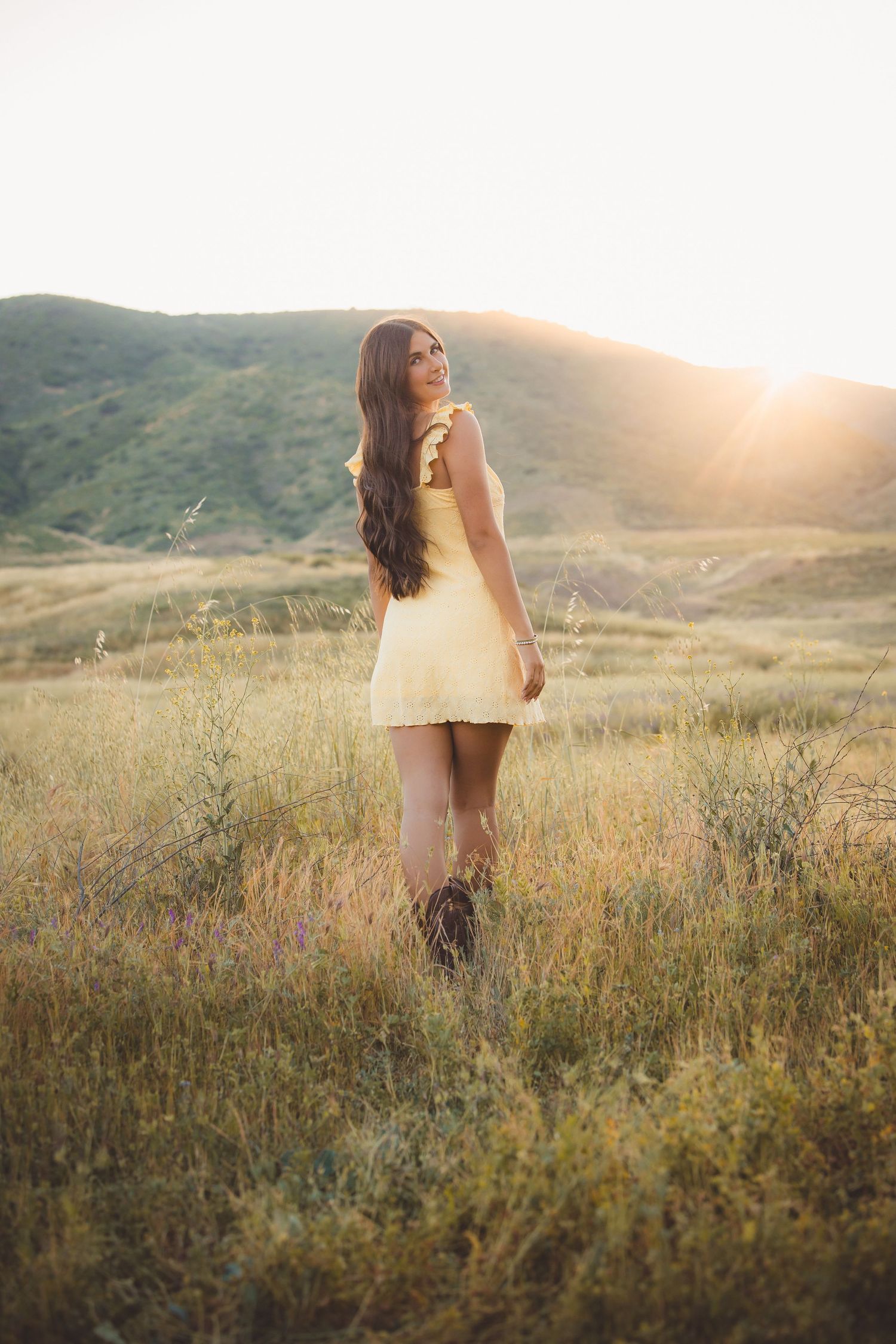 Young woman in yellow mini dress stands in sunlit meadow with green hills at golden hour.