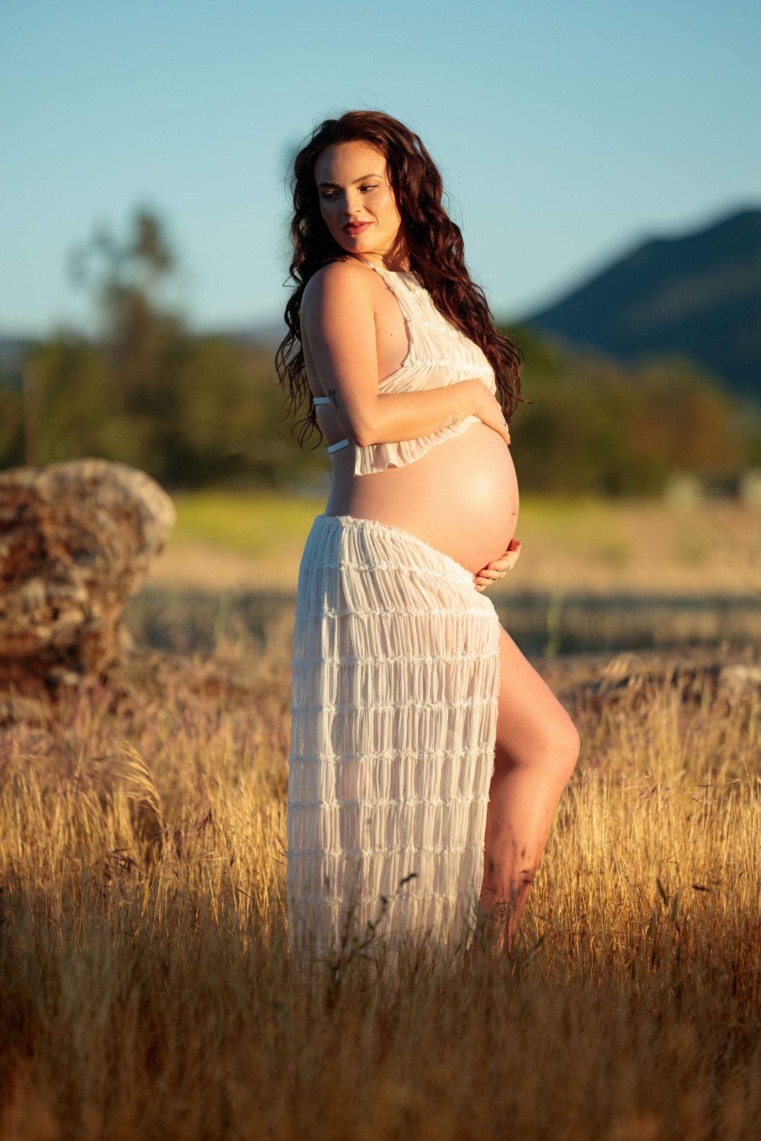 Pregnant woman in white dress standing in golden field with mountains in background during golden hour.