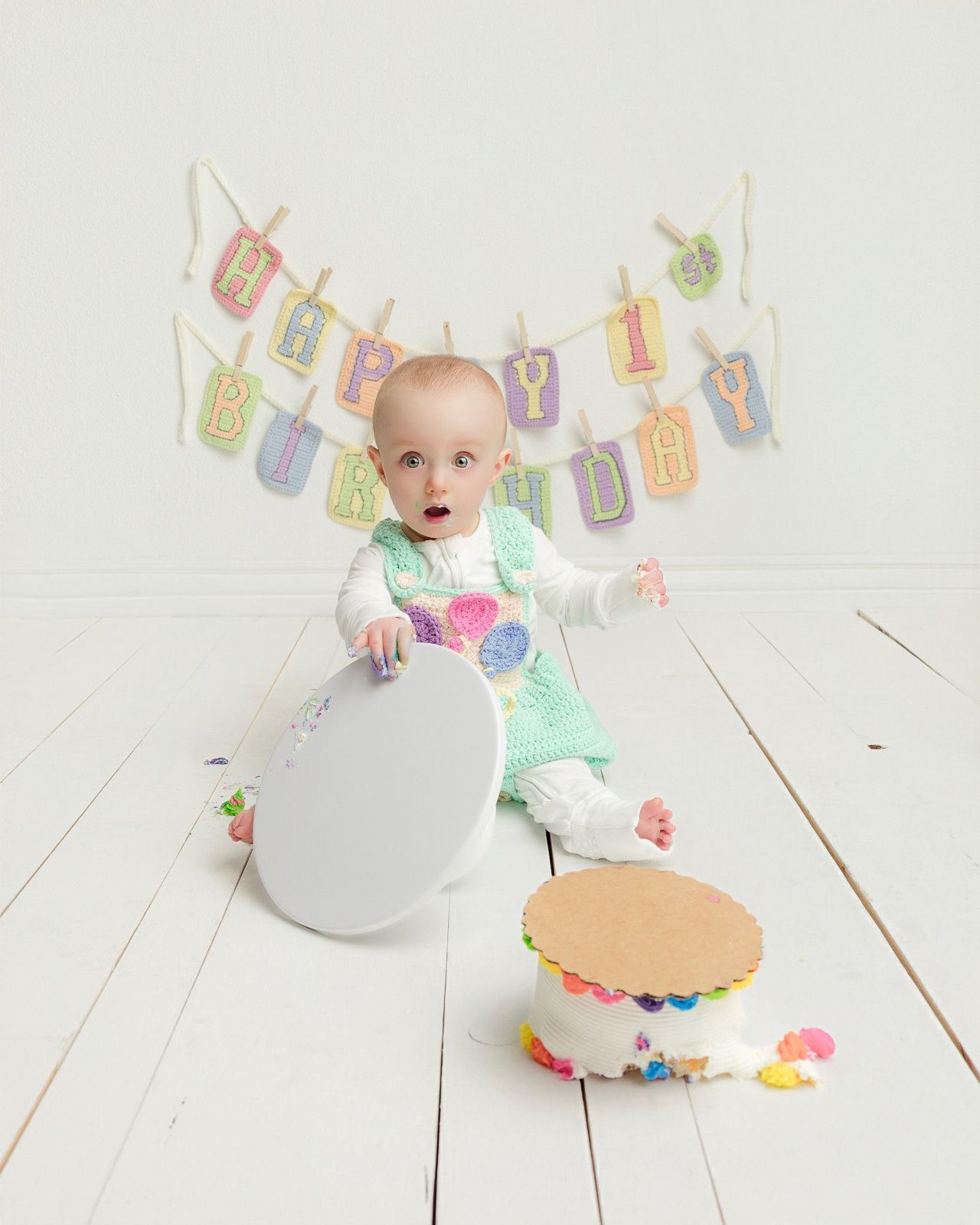 A surprised baby celebrates their first birthday with a smash cake and Happy Birthday banner on white wood floors.