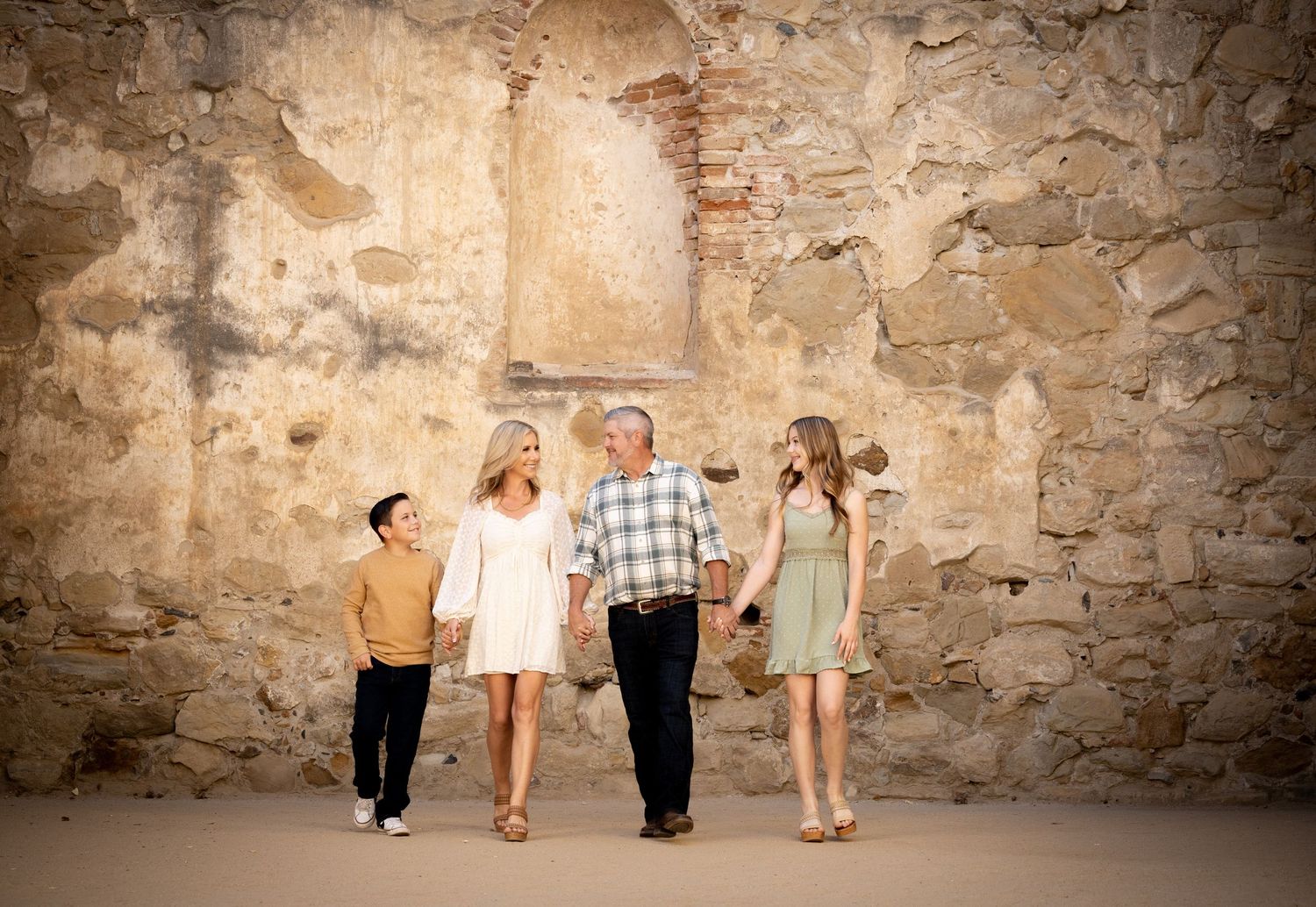 A family of four holding hands and walking together along a rustic stone wall in coordinated neutral outfits.