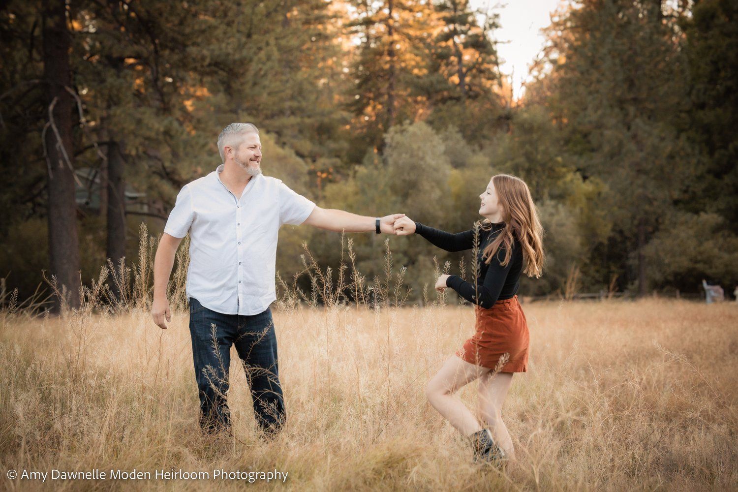 Joyful dancing moment captured in a golden field during sunset with evergreen trees in background.