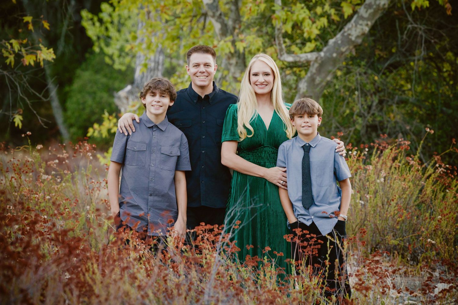 Smiling family of four posing outdoors in a field of autumn foliage, with two boys, a man in black and a woman in a green dress.