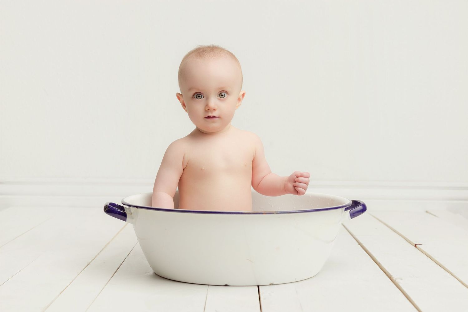Cute baby sitting in vintage white enamel tub on white wooden floor.