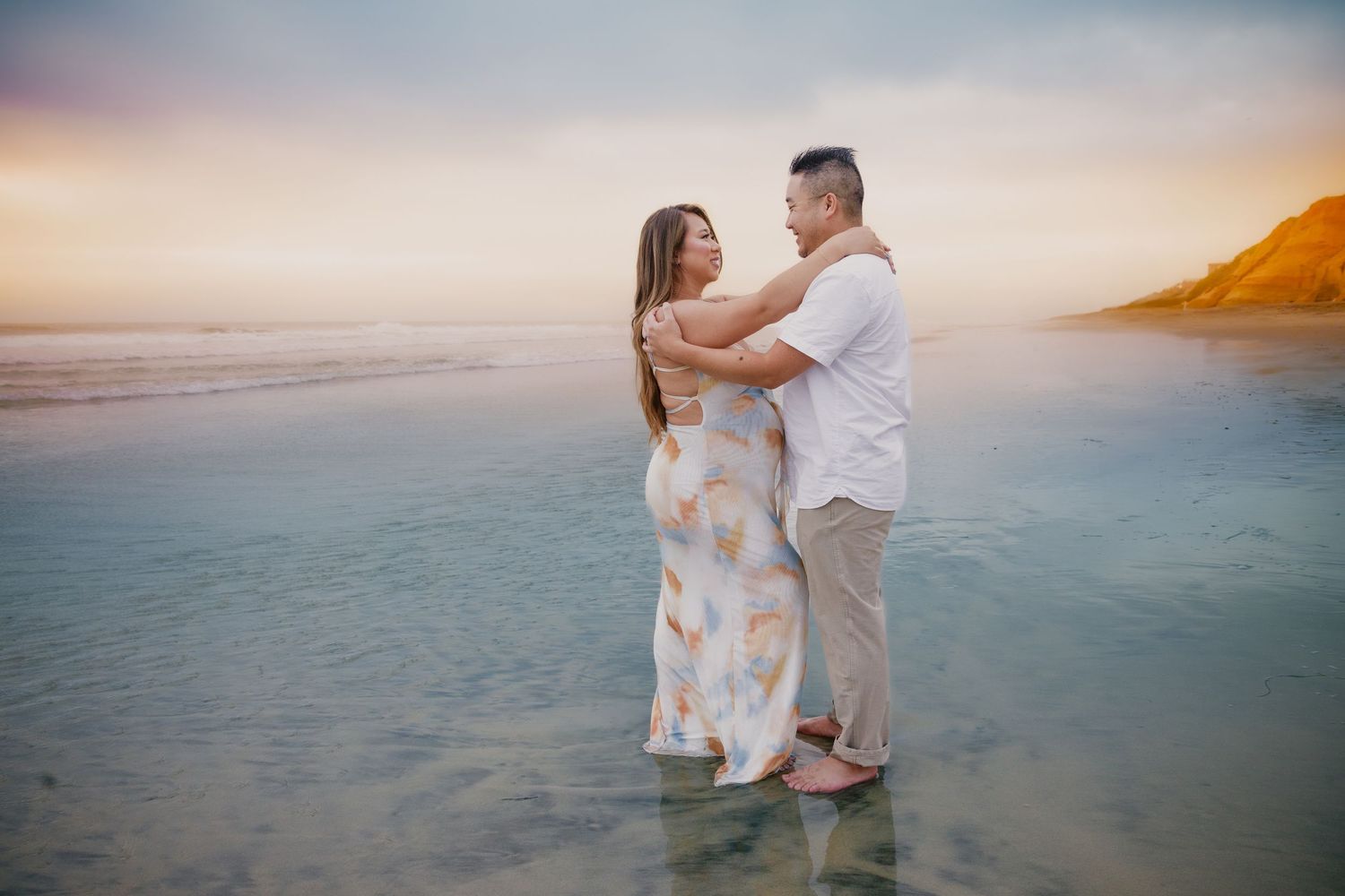 Couple embracing on a shallow beach at sunset, woman in floral dress and man in white shirt, ocean and colorful sky behind them.
