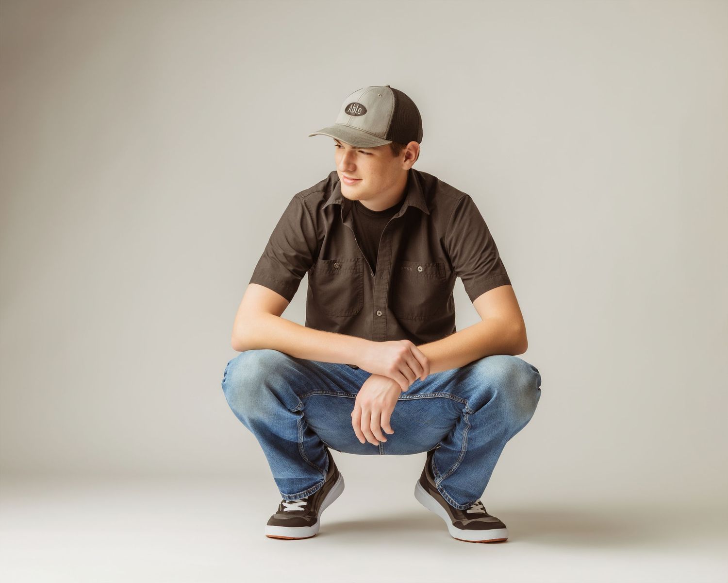 Young man in brown shirt, jeans, and trucker hat crouching low against a neutral gray studio background.