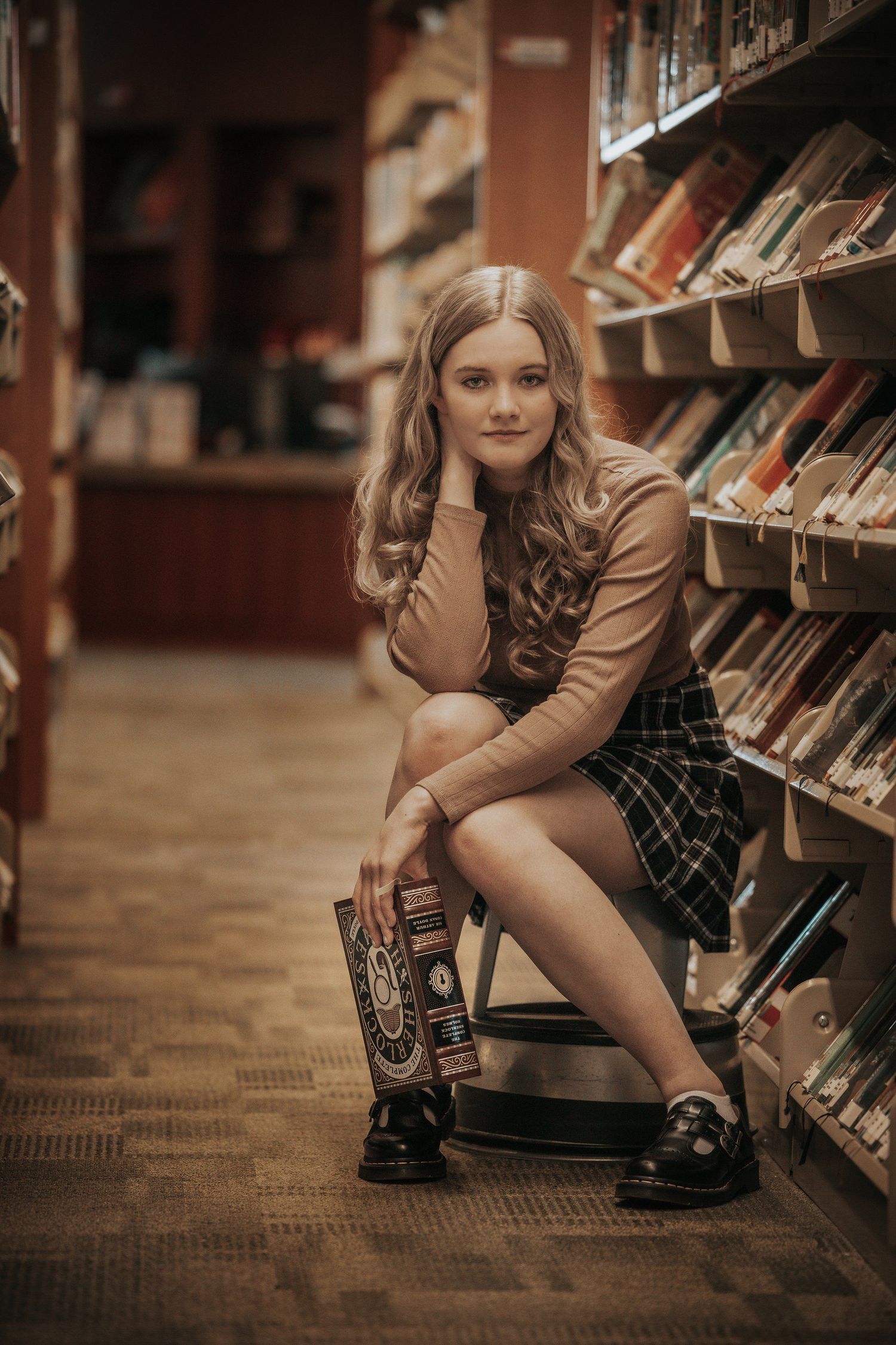 Young woman in plaid skirt sits between library bookshelves holding a book, wearing loafers in a warm-toned library.