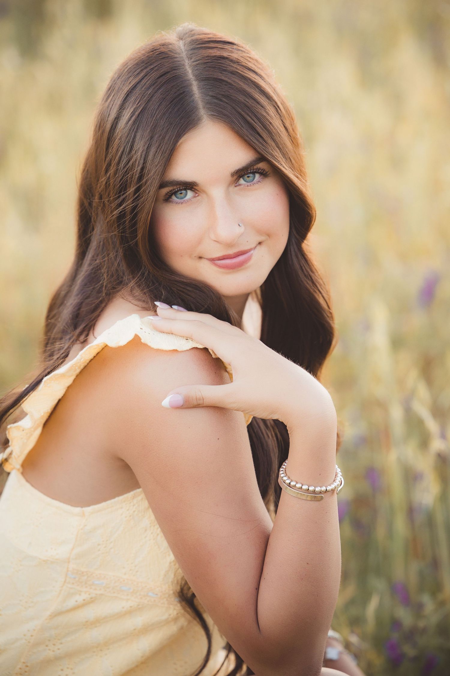 Young woman with long brown hair and blue eyes in yellow dress poses in golden field at sunset.