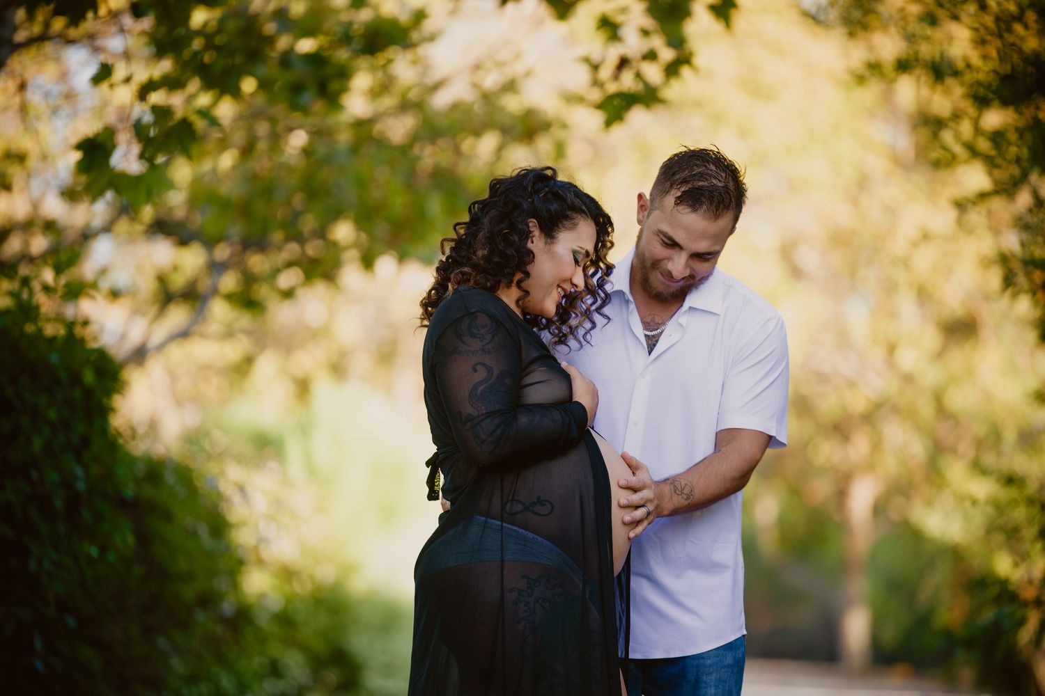 A couple shares a tender moment outdoors, the pregnant woman in a black dress embraced by her partner among lush green trees.