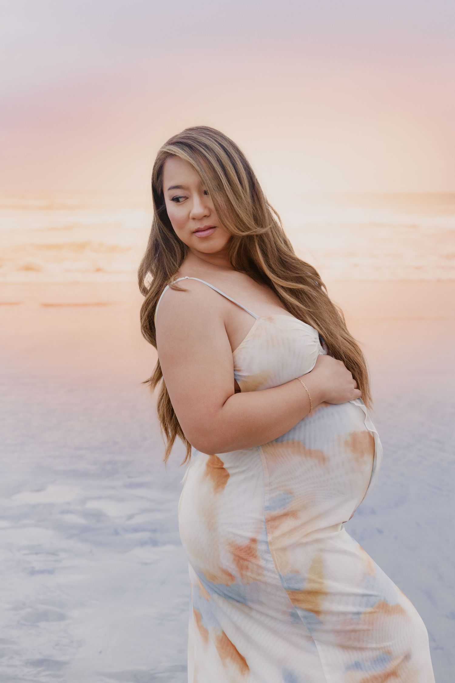 Pregnant woman in floral dress cradling baby bump at sunset beach, long hair flowing, soft pastel sky backdrop.