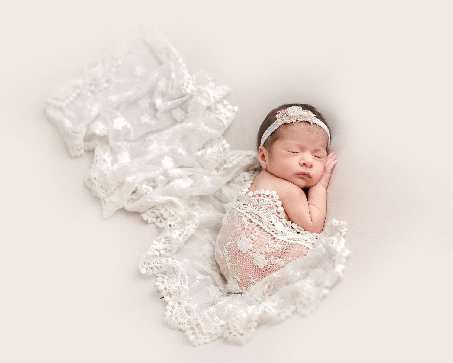 A sleeping newborn wrapped in delicate white lace fabric lies peacefully on a light pink background.