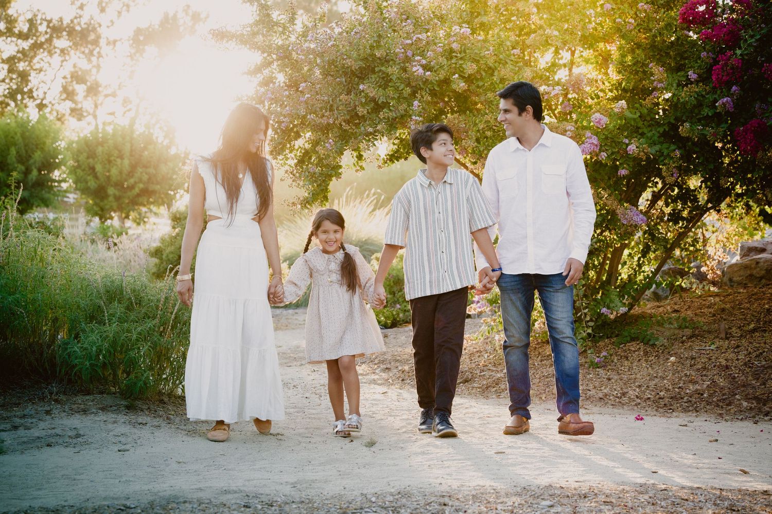 Connected family portraits in white outfits walking outdoors along a sunlit garden path at golden hour.