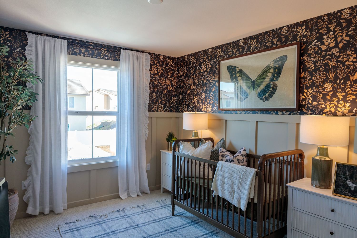 A nursery room with dark floral wallpaper features a white crib, dresser, and natural lighting from windows.