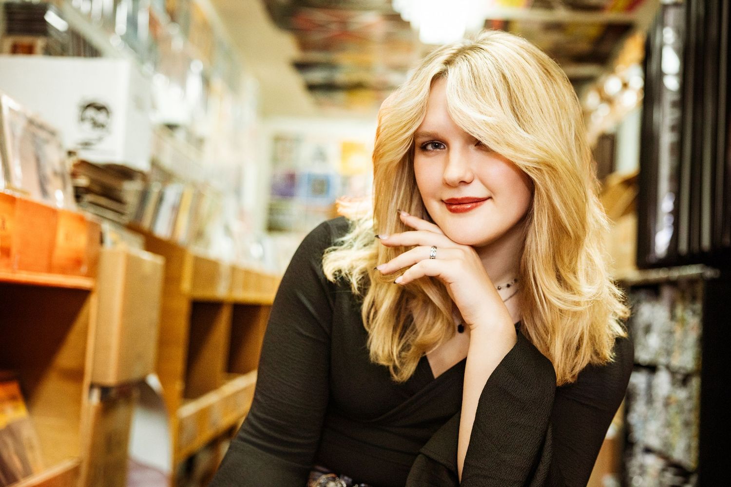 Blonde woman in black top posing in a bookstore, smiling with hand on chin.
