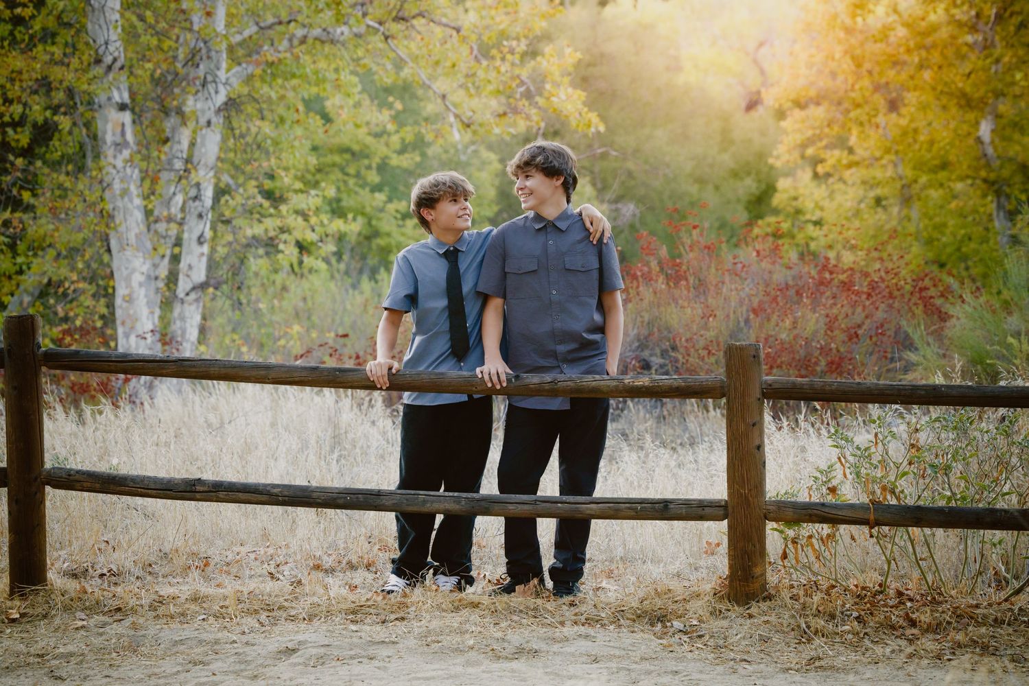 Two teenage boys in matching gray button-up shirts and dark pants lean on a rustic wooden fence with golden autumn trees behind them.