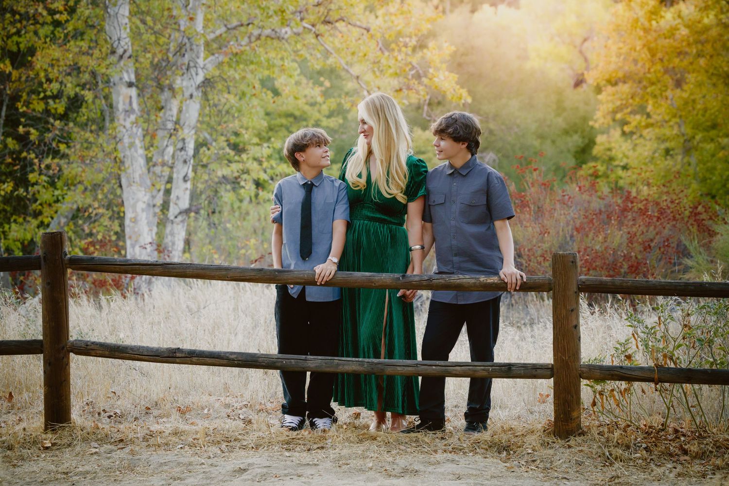 A woman in a green dress and two boys in blue shirts lean on a wooden rail fence in a sunlit autumn field with golden trees.