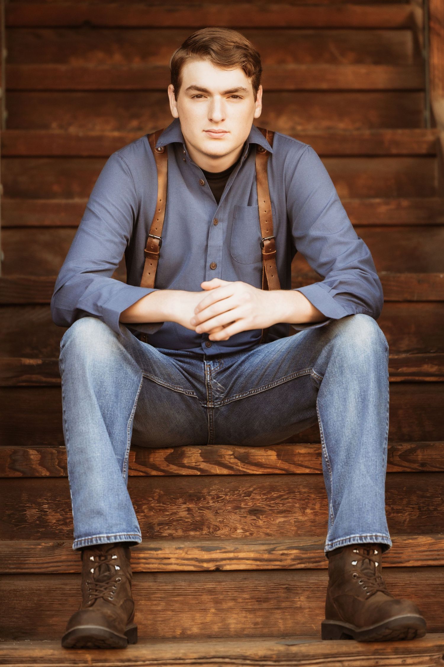 Young man in blue shirt and suspenders sitting on rustic wooden steps with boots.