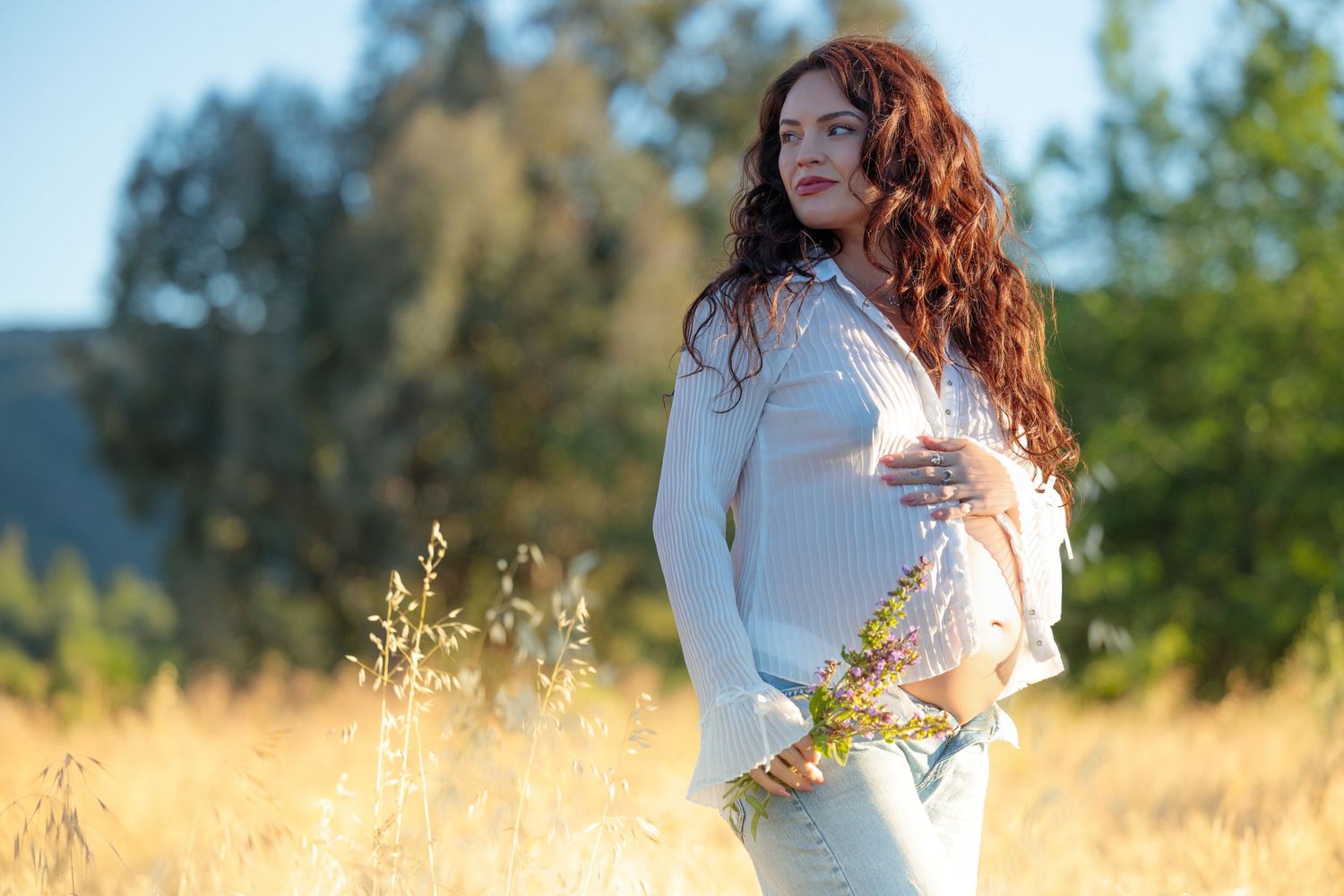 Pregnant woman with curly red hair in white sweater holding wildflowers in a sunny golden field.