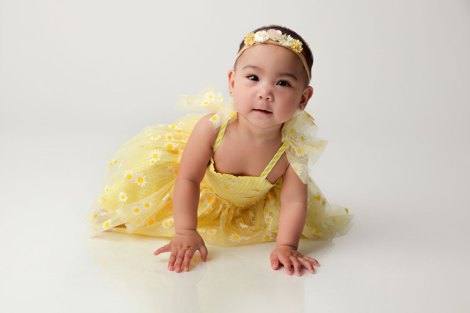 Baby girl in yellow tutu dress and floral headband crawling on white background.
