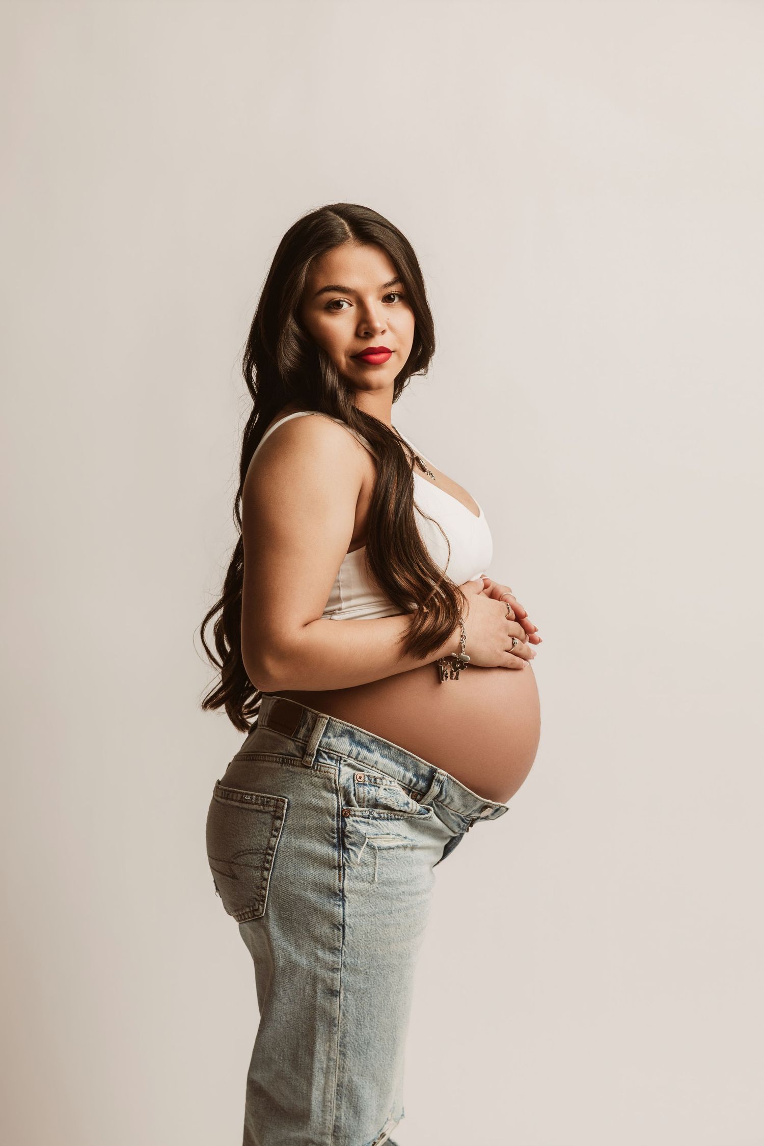 Maternity photo series showing pregnant figure in light denim overalls against neutral studio backdrop.