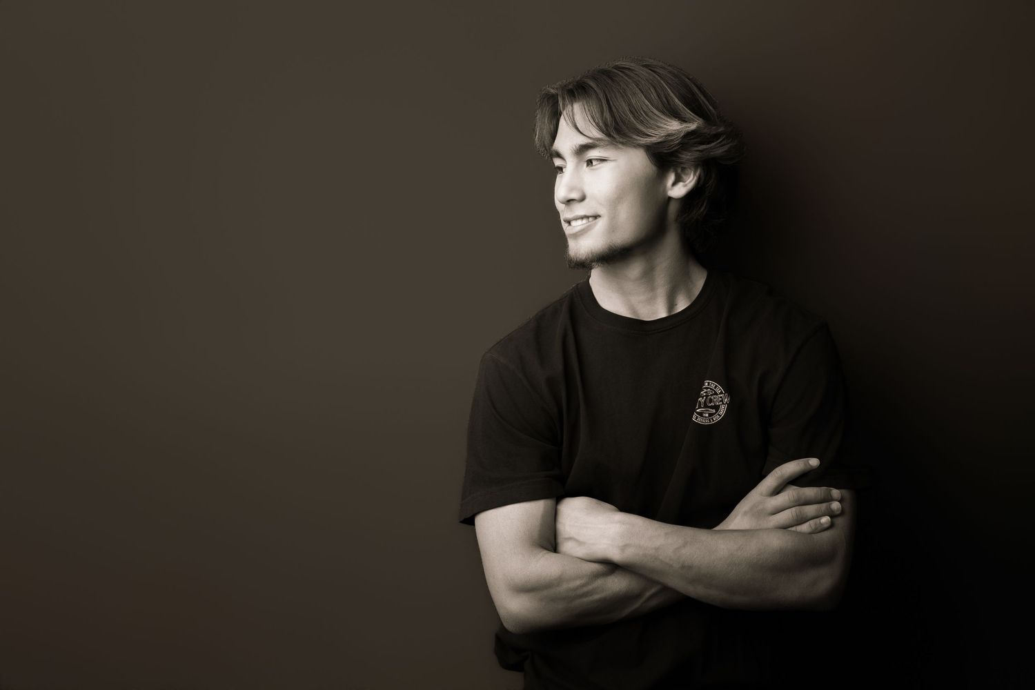Young man in black t-shirt posing confidently with arms crossed against dark background in monochrome portrait.