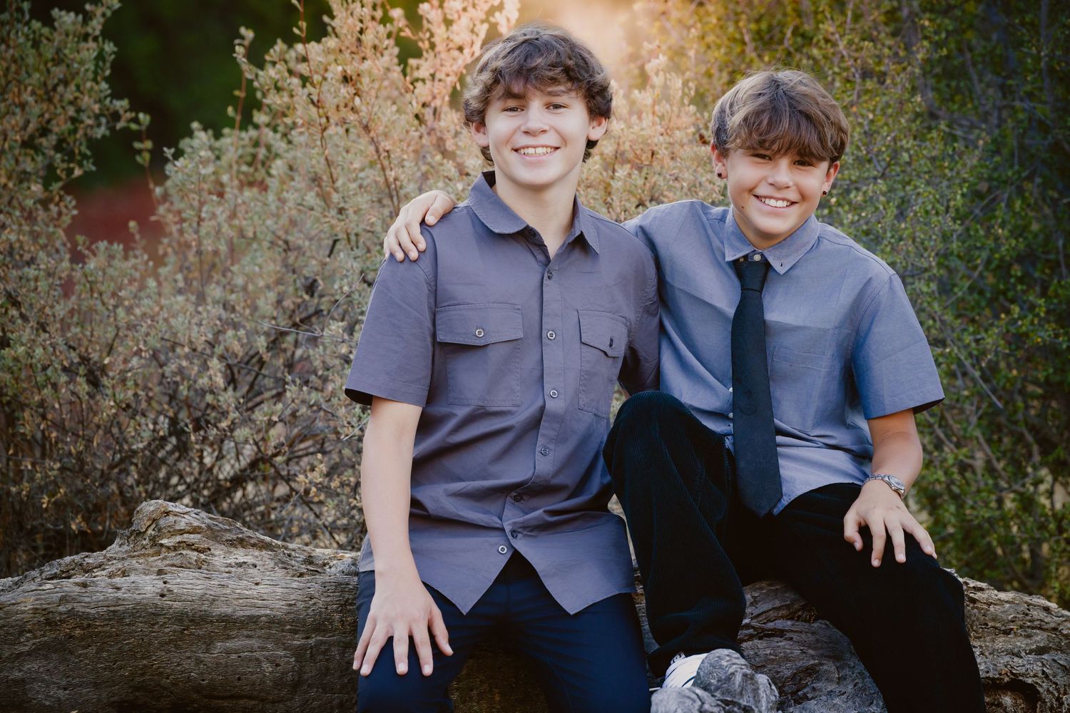 Two smiling boys in gray button-up shirts sit close together on a large log outdoors, with golden autumn shrubs and trees behind them.