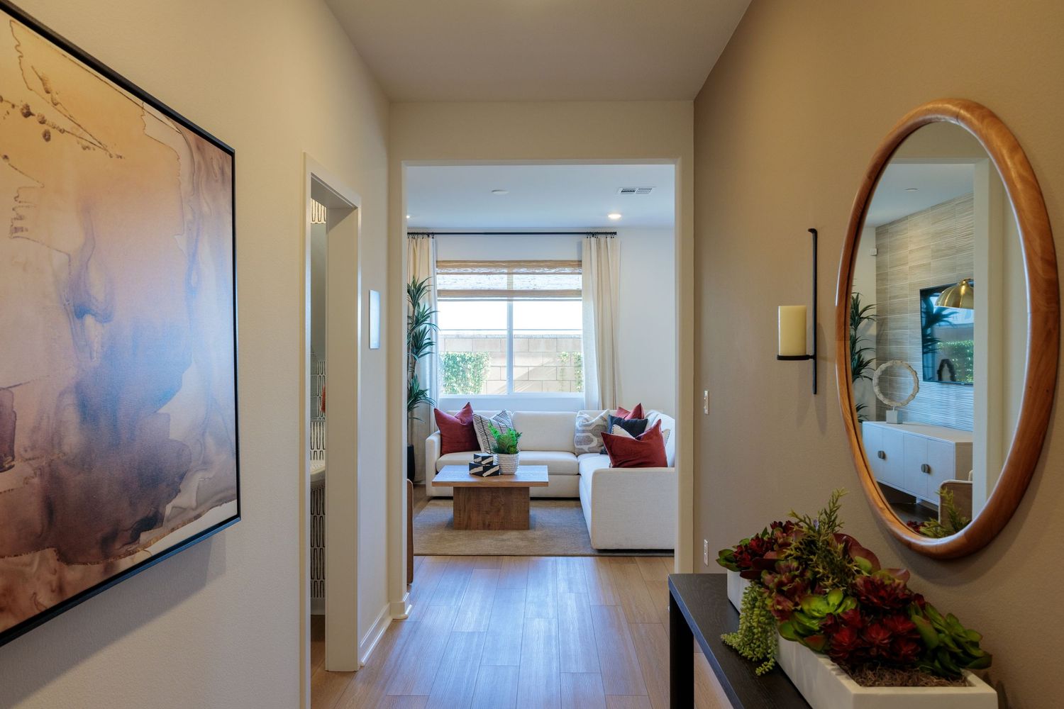 Modern hallway with hardwood floors leads to bright living room featuring round mirror, artwork and window seating area.
