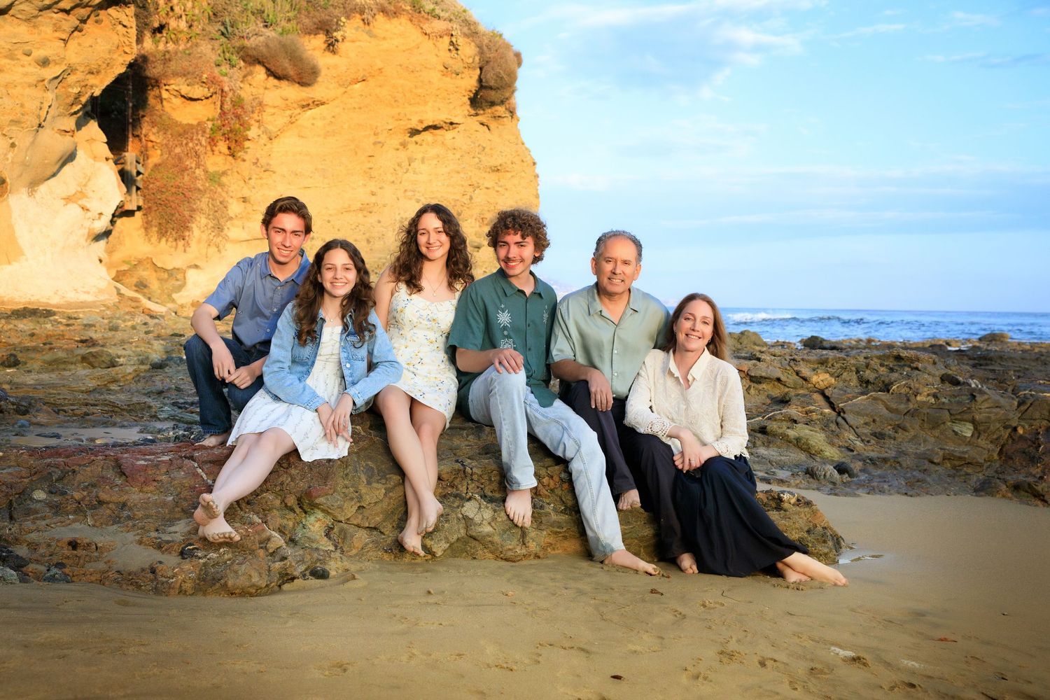 Family in casual attire relaxing and posing together at a scenic beach with cliffs and rocks at sunset.