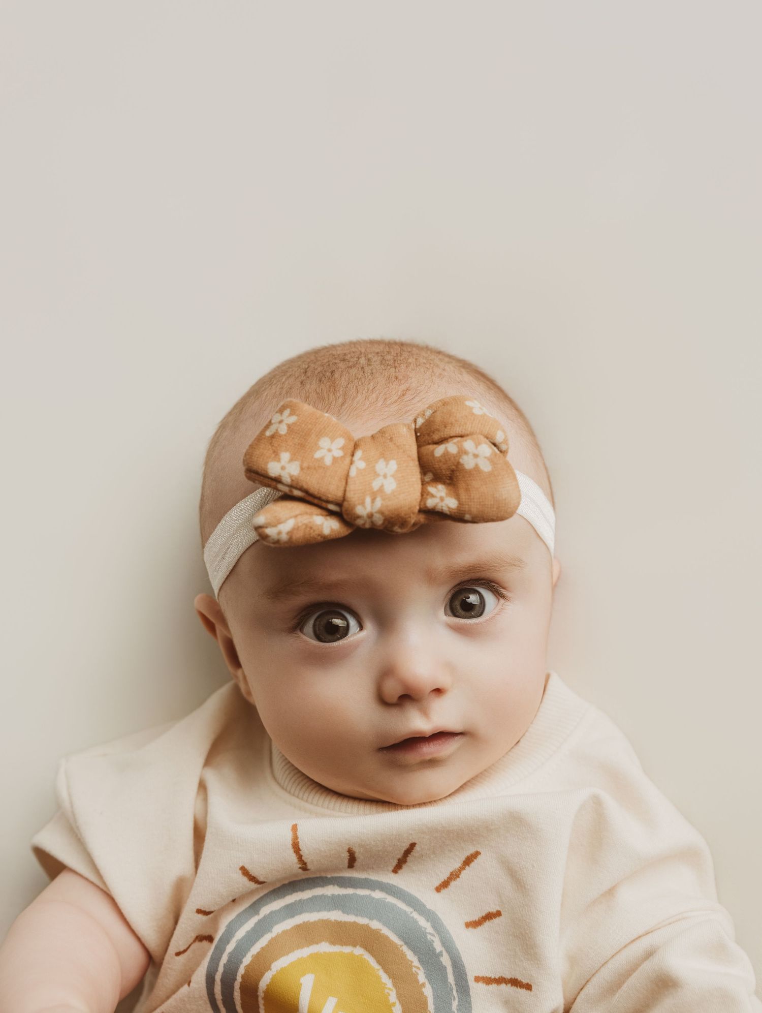 A baby wearing a cream-colored outfit looks curiously at the camera with wide eyes and a slight grin.