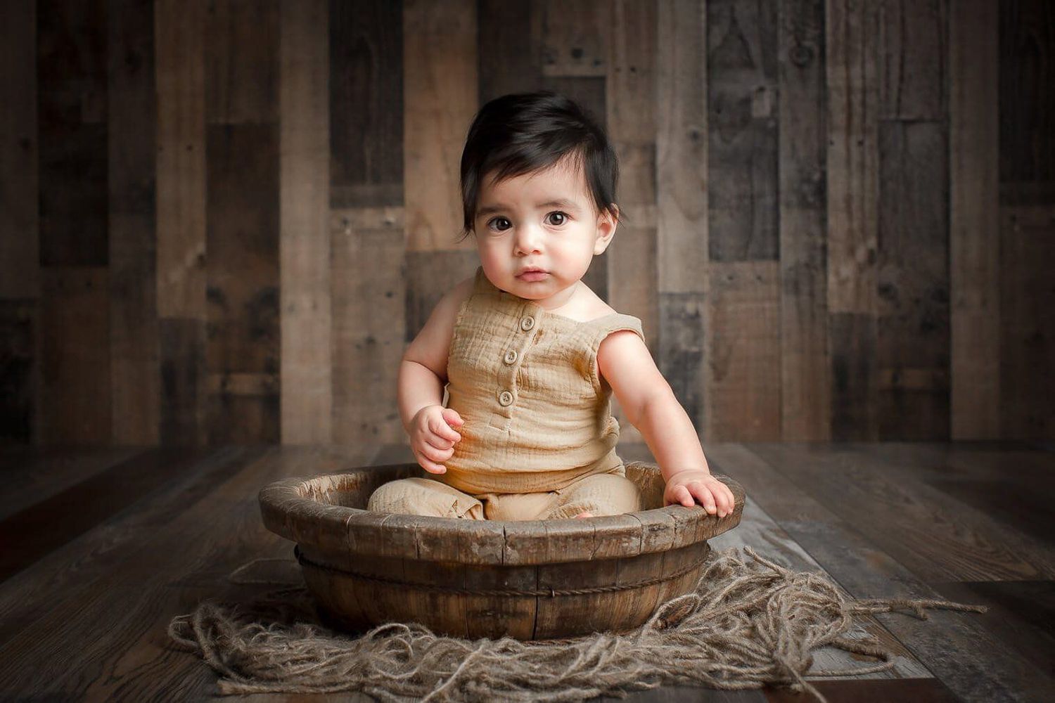 A cute baby in a beige romper sits in a rustic wooden bowl on a wood plank background.