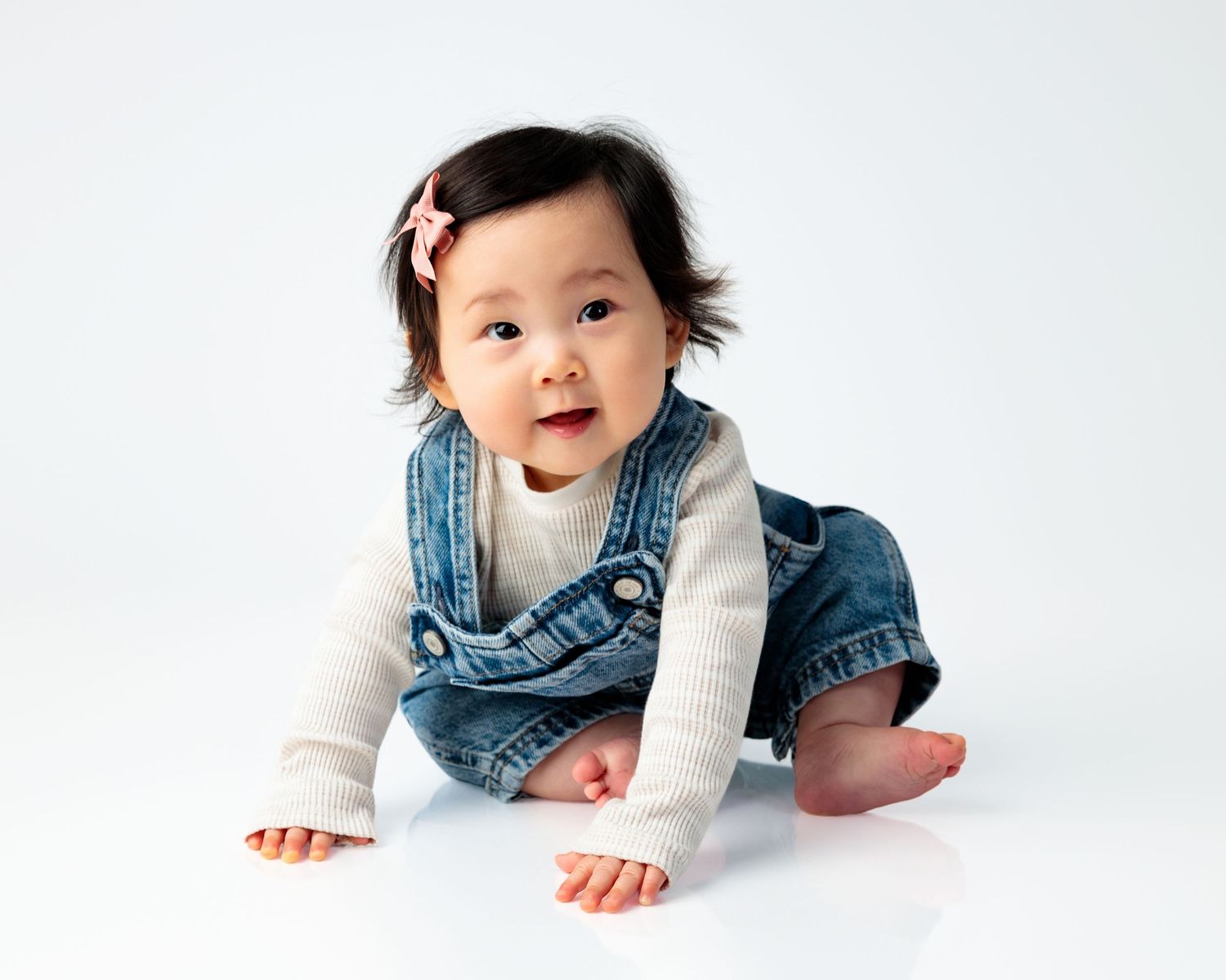 A baby in a denim vest lies on their stomach on a white background with a bright smile.