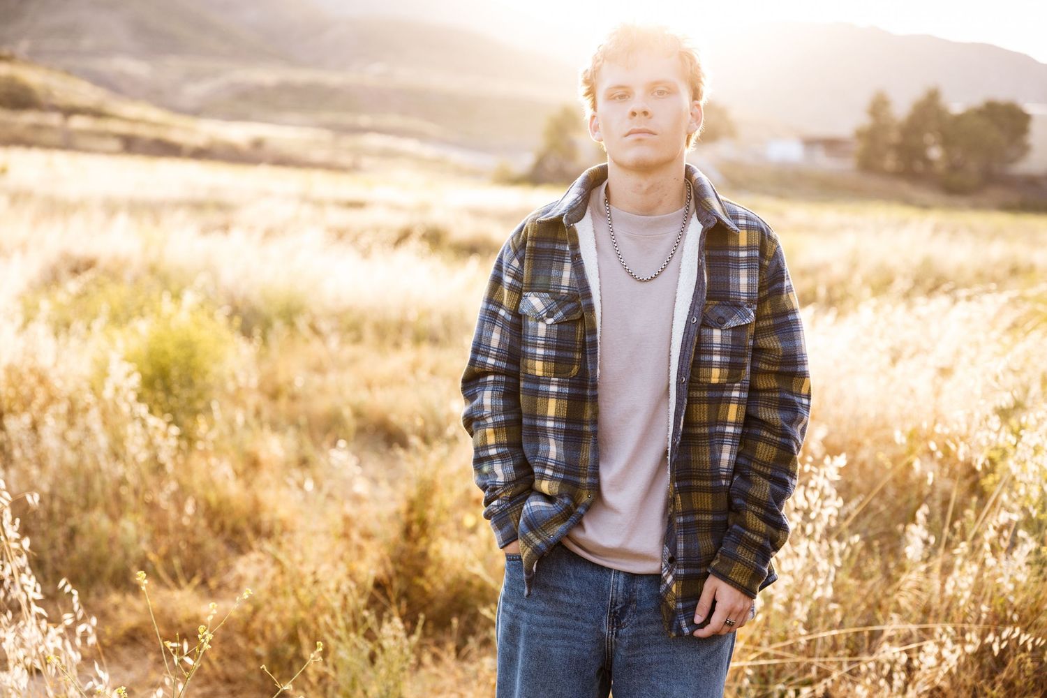 Young man in plaid flannel jacket and jeans standing in golden sunlit field outdoors.