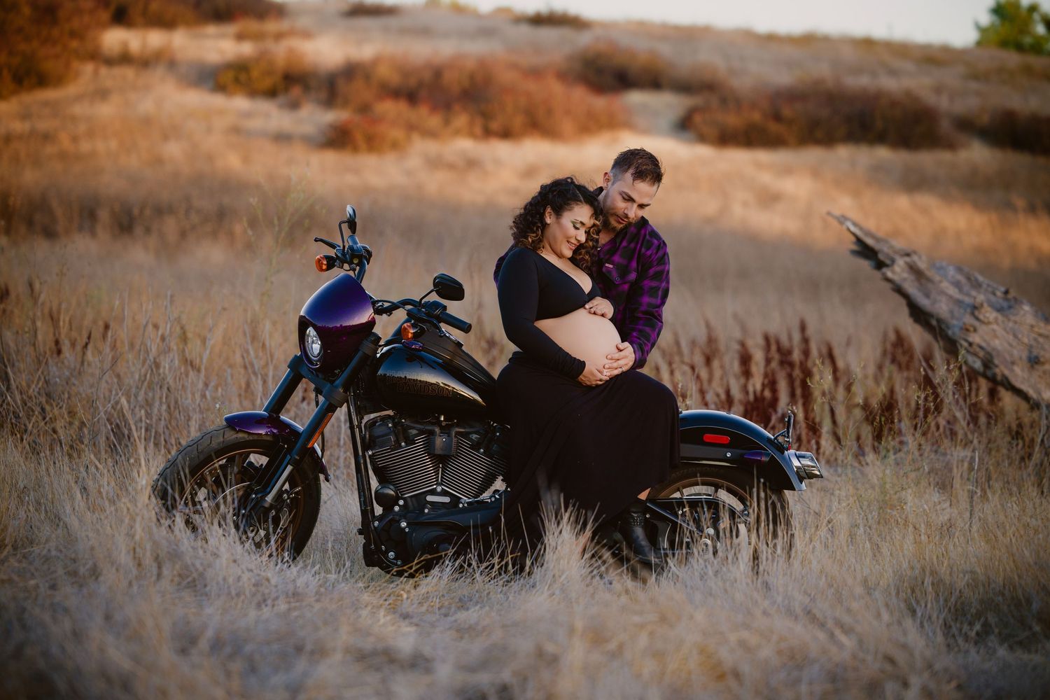Expecting couple embraces on a motorcycle in a golden field at sunset, celebrating their pregnancy outdoors.