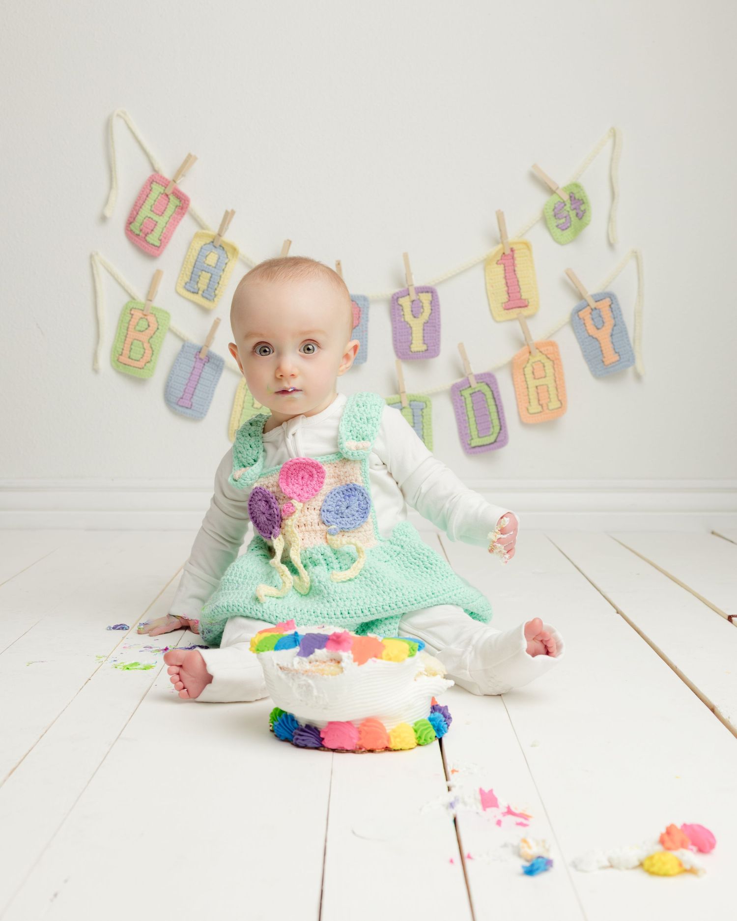 Baby in mint green dress plays with colorful yarn balls on white wooden floor while alphabet letters hang above.