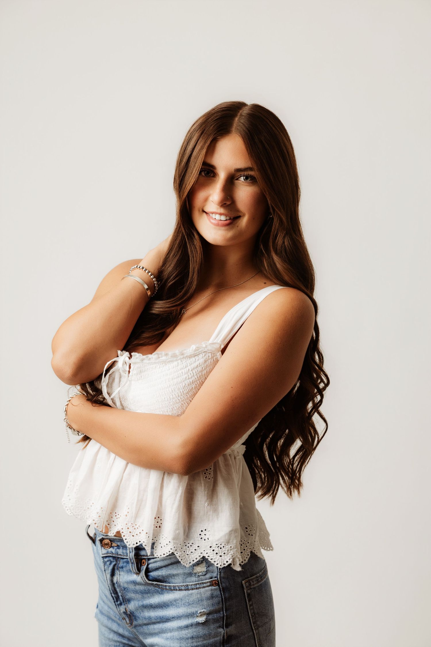 Young woman with long brown hair smiling in white eyelet tank top and jeans against light background.