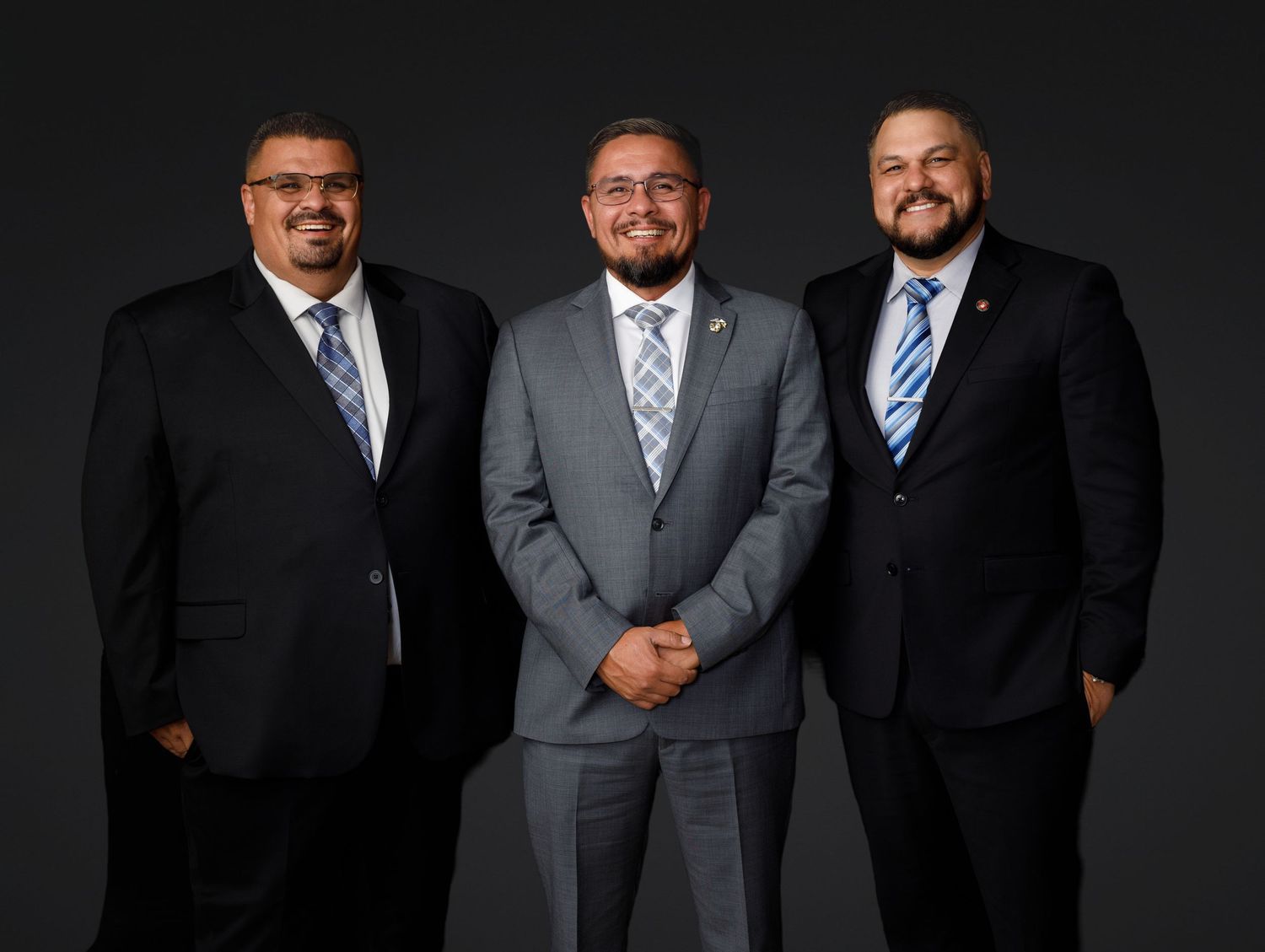 Three businessmen in suits and ties stand together for a professional portrait against a dark background.
