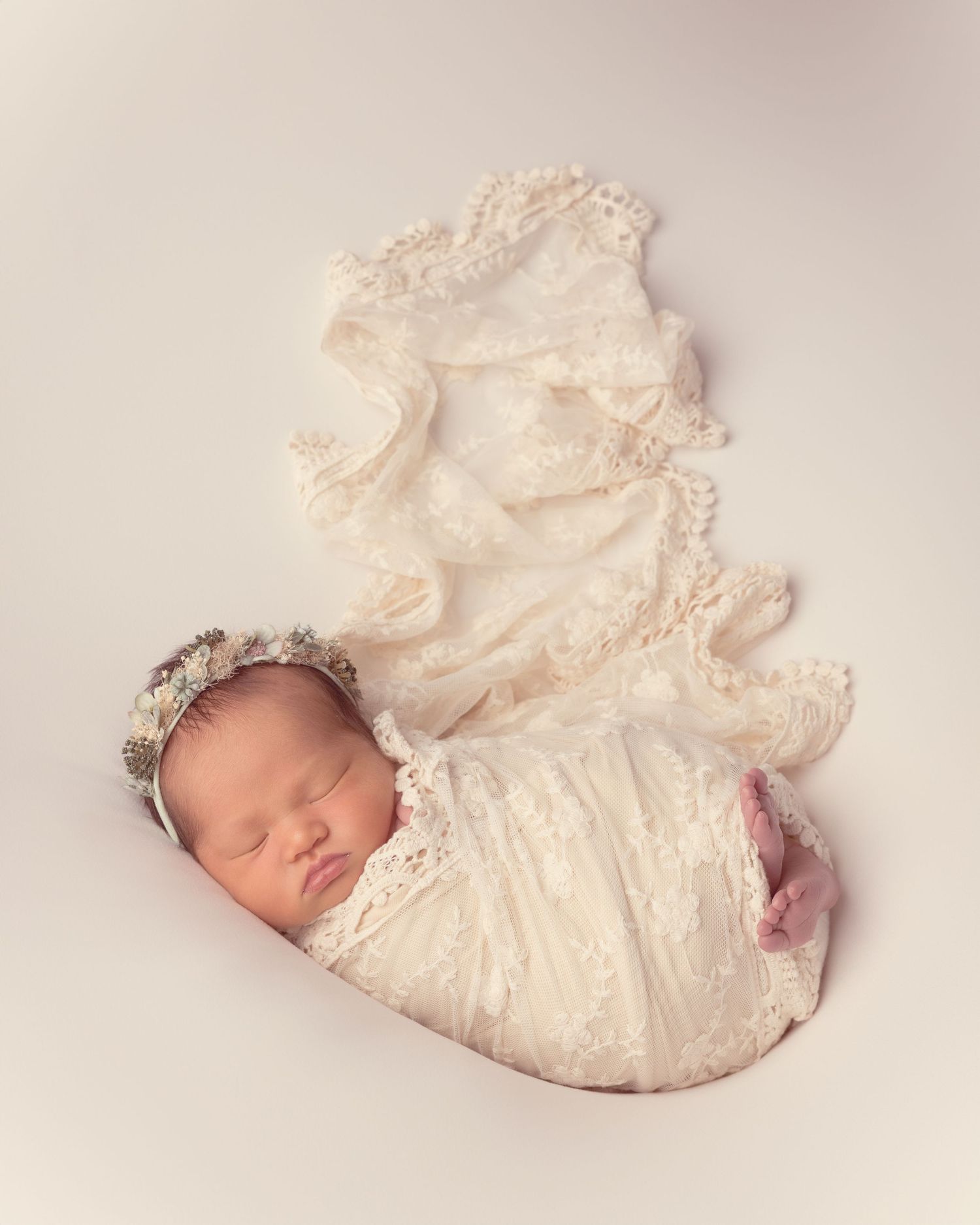 A sleeping baby in a white lace dress lies on a soft cream backdrop in a delicate newborn portrait.