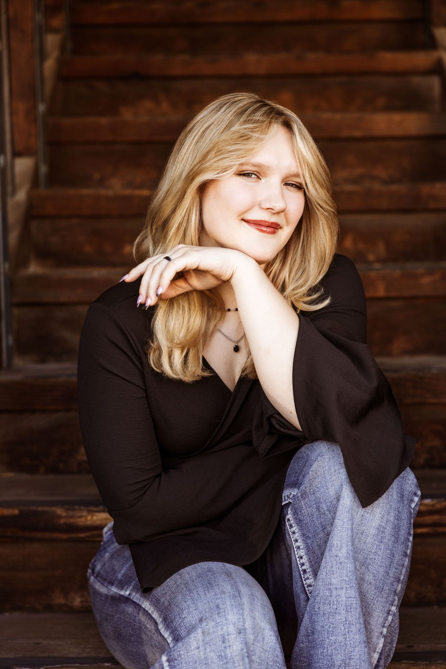 A person in a black top and jeans sits casually against a rustic wooden wall background.