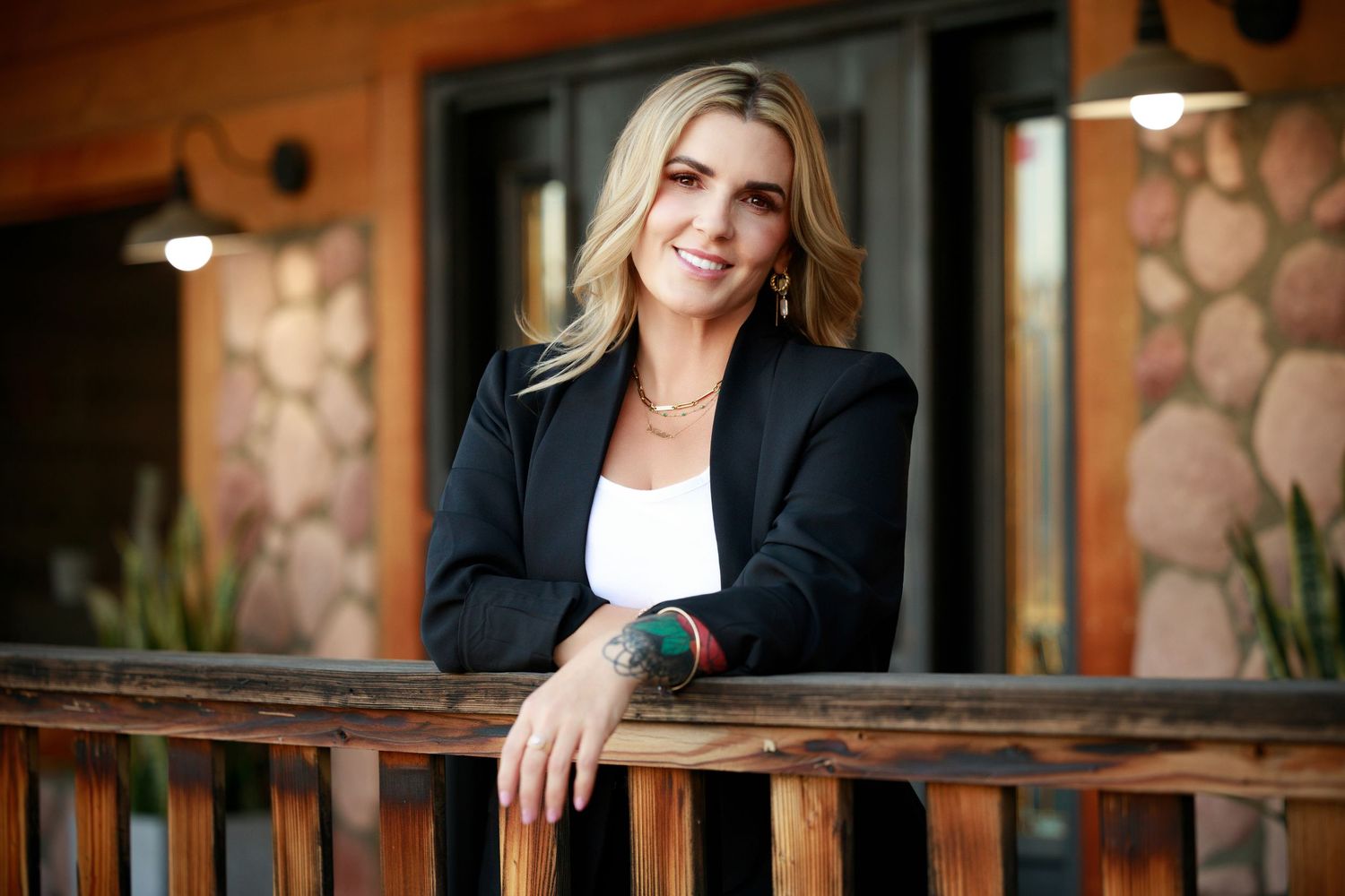 Smiling blonde woman in black blazer and white top leaning on wooden railing outside a rustic building.