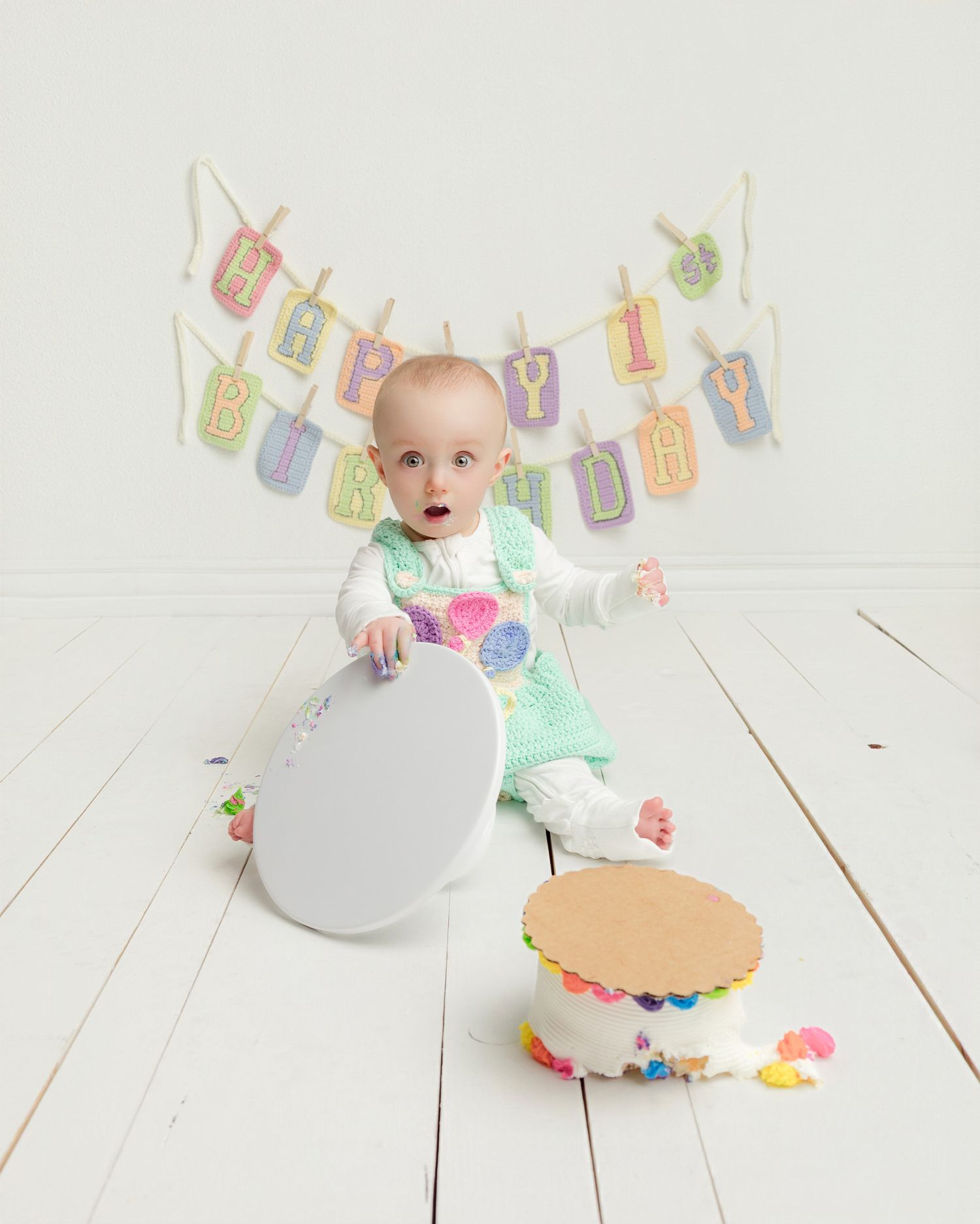 Baby girl in green outfit sits beside smashed birthday cake under Happy Birthday banner on white floor.